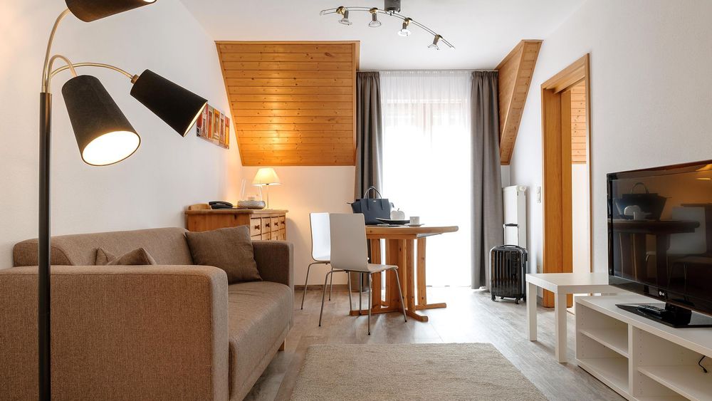 Wohnbereich mit braunem Sofa, Stehlampe, Esstisch mit zwei Stühlen und Fernseher.