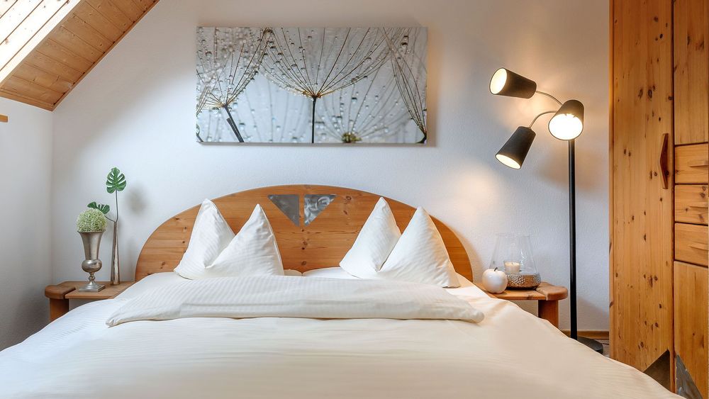 Schlafzimmer mit Holzbett, weißer Bettwäsche und Wandbild mit Makroaufnahme von Pusteblumen.