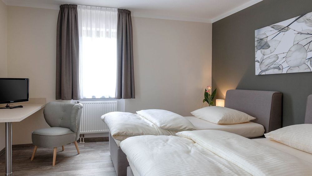 Doppelzimmer mit zwei Betten, grauer Wand, grauem Sessel und Fernseher auf hellem Schreibtisch.