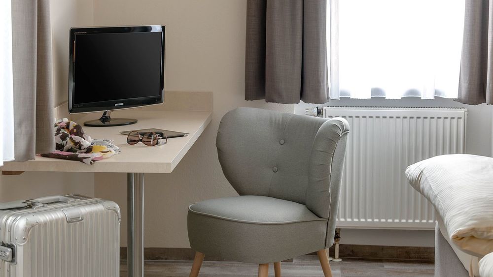 Schreibtisch mit grauem Stuhl, Fernseher und Koffer neben Bett mit hellen Bettbezügen.