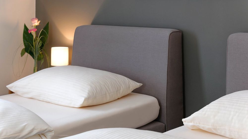 Nahaufnahme eines Bettes mit grauem Kopfteil und Nachttischlampe, Blume im Hintergrund.
