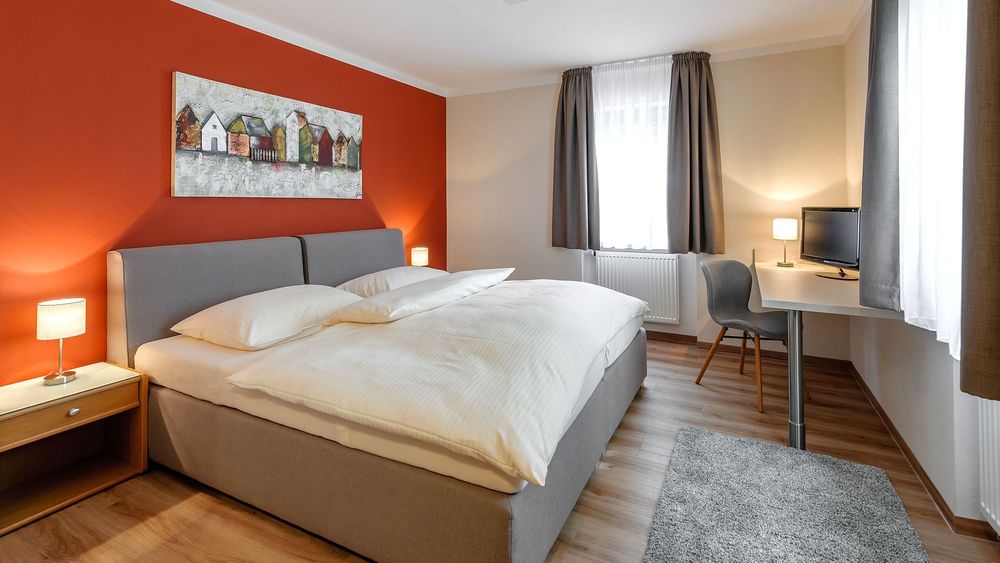 Helles Doppelzimmer mit roter Wand, grauem Bett und Schreibtisch mit Fernseher am Fenster.