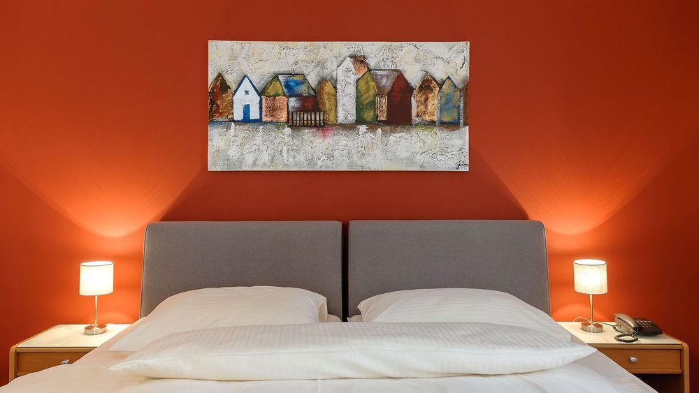 Doppelbett mit grauem Kopfteil und roter Wand, beleuchtet von zwei Nachttischlampen.