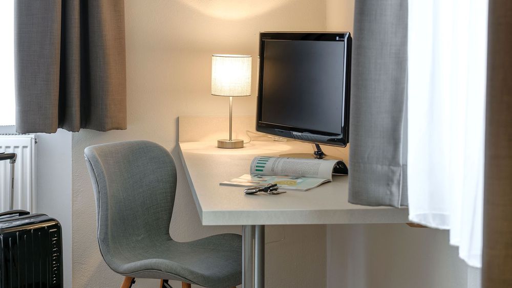 Schreibtisch mit Fernseher, Lampe und grauem Stuhl neben Fenster mit grauen Vorhängen.