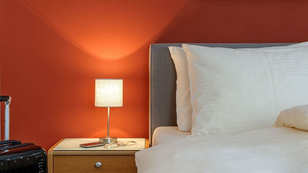 Doppelbett mit Nachttischlampe, roter Wand und Koffer am Bettrand.