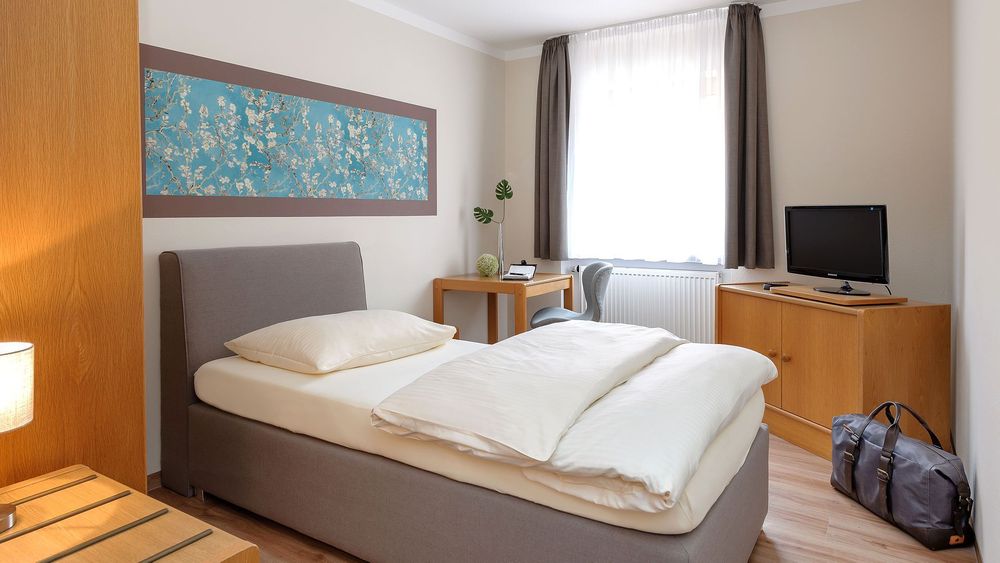 Einzelzimmer mit grauem Bett, hellem Holzmobiliar und Sporttasche am Boden neben dem Schrank.